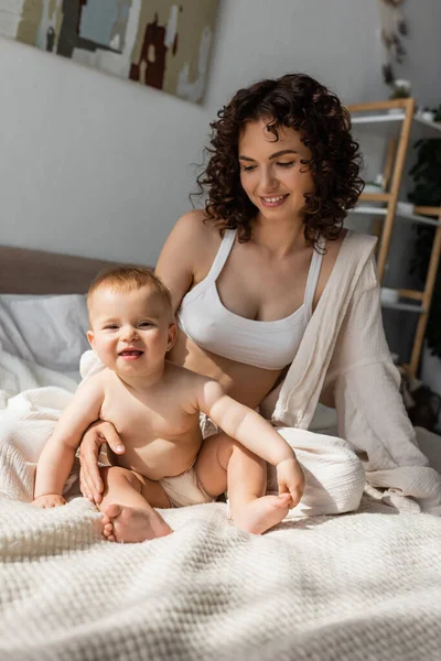 Щаслива мати в лаунж-одязі з верхньою частиною врожаю, що сидить поруч з веселою дочкою-немовлям у спальні — стокове фото