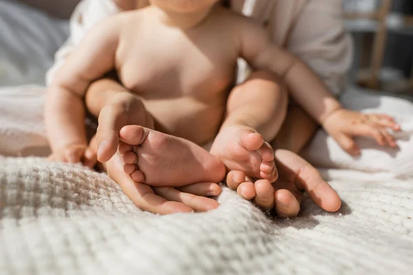 Vista recortada de la madre sosteniendo en las manos pequeños pies desnudos de la hija lactante en el dormitorio — Stock Photo
