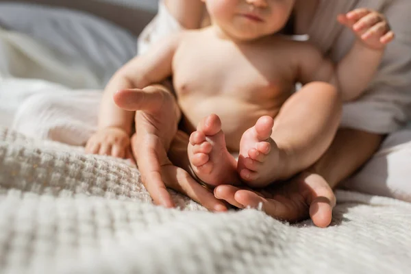 Partial view of mother holding in hands tiny bare feet of infant daughter in bedroom — Stock Photo