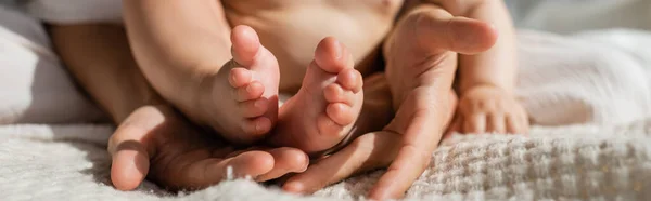 Vista recortada de la madre sosteniendo en las manos pequeños pies desnudos de la hija lactante en el dormitorio, pancarta - foto de stock