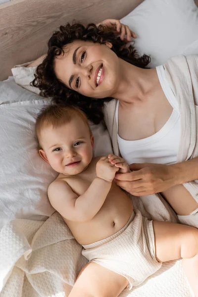 Vista superior de la madre rizada complacida en ropa de salón acostada con la niña en la cama - foto de stock