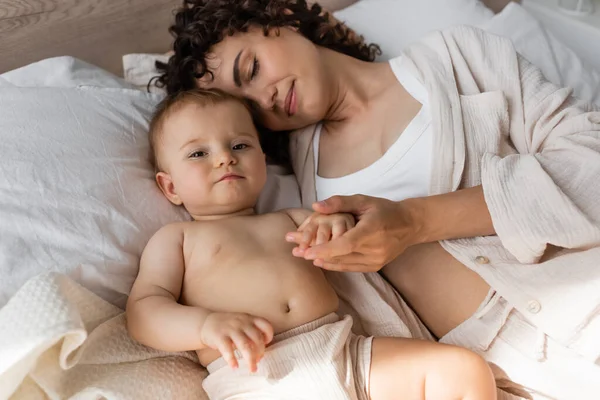 Высокий угол зрения кудрявой матери в пеленках, лежащей с маленькой девочкой на кровати — стоковое фото