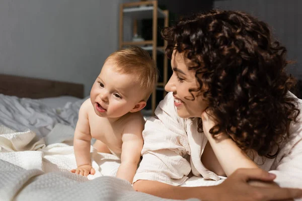 Glückliche Mutter mit lockigem Haar schaut neugieriges Mädchen auf dem Bett an — Stockfoto