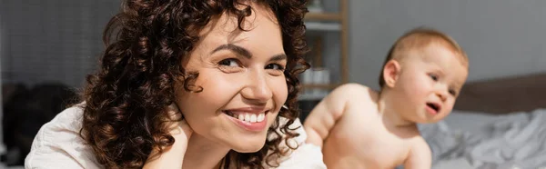 Happy mother with curly hair looking at camera near blurred baby girl, banner — Stock Photo