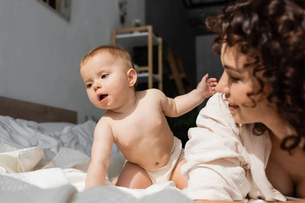 Mignon bébé fille assis sur le lit près floue et frisée mère — Photo de stock
