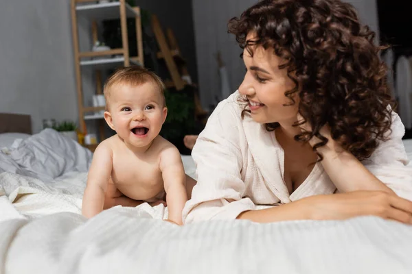 Fröhliche Mutter mit lockigem Haar schaut süße kleine Tochter auf dem Bett an — Stockfoto