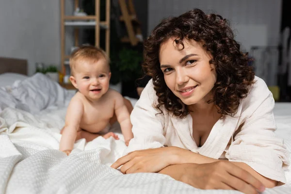 Cheerful mother with curly hair looking at camera joyful baby daughter on bed — Stock Photo