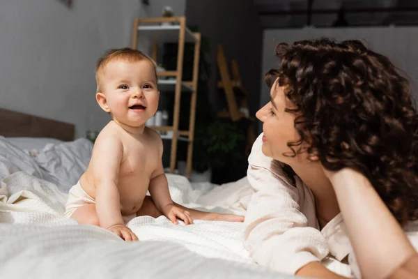 Mother with curly hair looking at happy baby daughter sitting on bed — Stock Photo