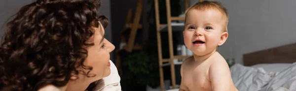 Mère avec les cheveux bouclés regardant la fille heureuse de bébé dans la chambre, bannière — Photo de stock