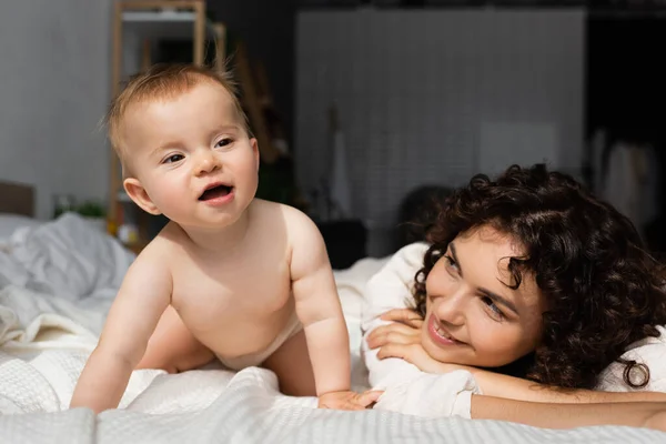 Cheerful mother with curly hair looking at curious baby daughter crawling on bed — Stock Photo
