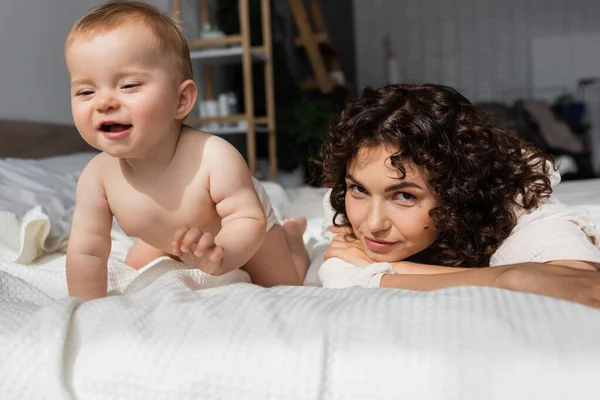 Frau mit lockigem Haar blickt in Kamera neben neugieriger Baby-Tochter, die auf Bett kriecht — Stockfoto