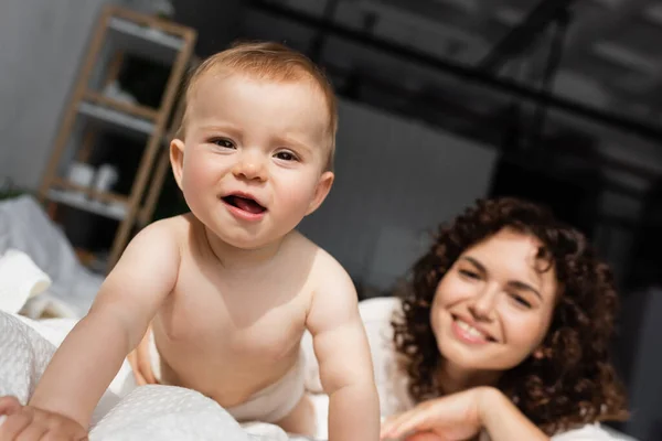 Babymädchen krabbelt auf Bett und blickt in Kamera in der Nähe verschwommene Mutter mit lockigem Haar — Stockfoto