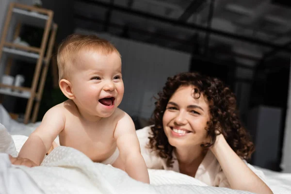 Aufgeregtes Säuglingsmädchen krabbelt auf Bett neben verschwommener Mutter mit lockigem Haar — Stockfoto