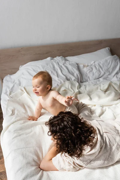 High angle view of happy infant girl crawling near mother with curly hair lying on bed — Stock Photo