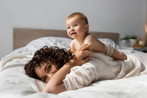 Cheerful infant baby lying on back of curly mother in bedroom — Stock Photo