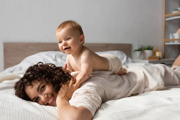 Cheerful infant baby lying on back of curly and happy mother in bedroom — Stock Photo