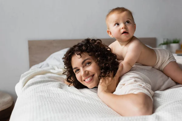 Bambino neonato sdraiato sul retro della madre felice con i capelli ricci in camera da letto — Foto stock