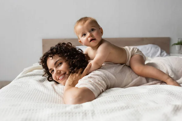Infant child lying on back of happy mother with curly hair in bedroom — Stock Photo
