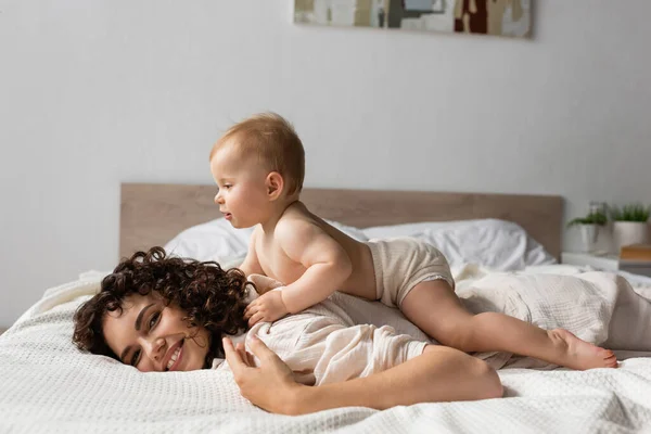 Cheerful infant baby lying on back of curly mother smiling in bedroom — Stock Photo