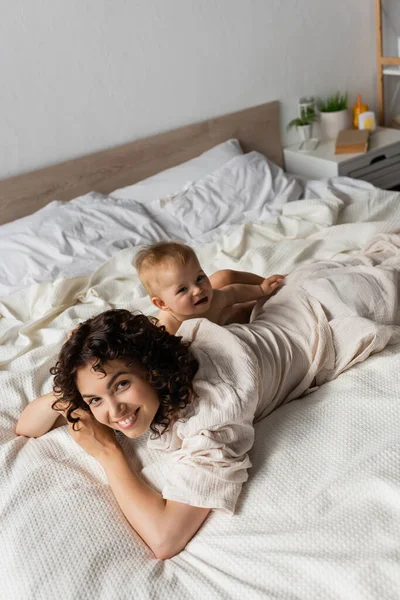 High angle view of infant baby lying near cheerful and curly mother in bedroom — Stock Photo