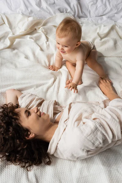 Вид сверху кудрявой матери, улыбающейся, глядя на веселую младенческую дочь на кровати — стоковое фото