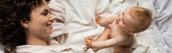 Vista superior da mãe encaracolada sorrindo enquanto olha para a filha infantil alegre na cama, banner — Fotografia de Stock