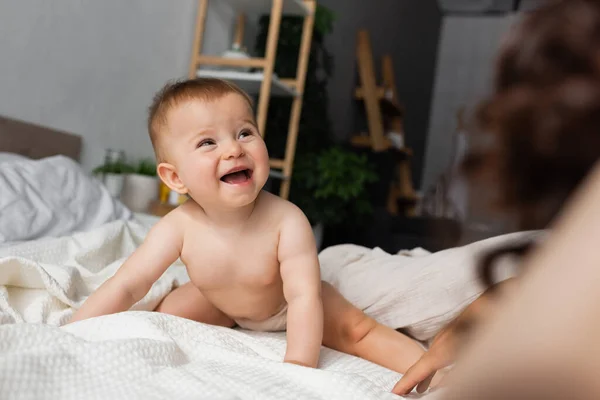 Happy infant baby sitting on bed and looking at blurred mother in bedroom — Stock Photo