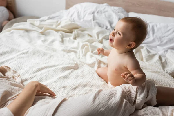 Happy infant baby sitting on bed and looking at mother resting in bedroom — Stock Photo