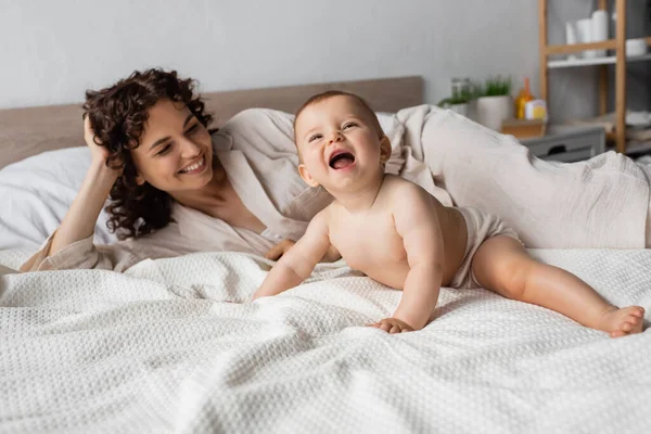 Glückliche und lockige Mutter lächelt, während sie aufgeregte Säuglinge auf dem Bett ansieht — Stockfoto