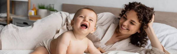 Feliz y rizada madre sonriendo mientras mira emocionada hija lactante en la cama, pancarta - foto de stock