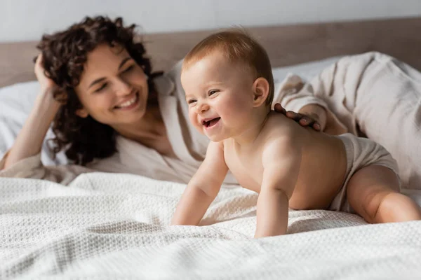 Madre borrosa y feliz sonriendo mientras mira a la niña alegre en la cama - foto de stock