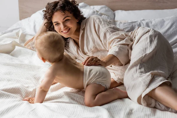 Mãe feliz com cabelo encaracolado sorrindo enquanto olha para a menina infantil rastejando na cama — Fotografia de Stock