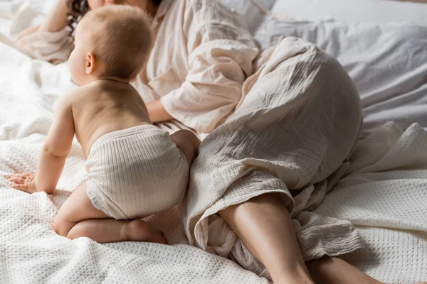 Vista recortada de la madre en ropa de salón acostado cerca de la hija lactante en la cama - foto de stock