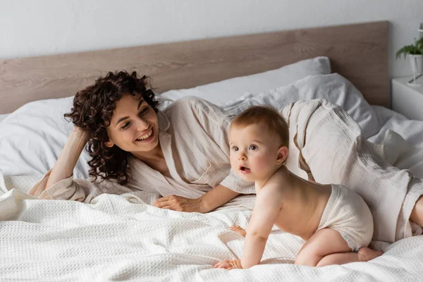 Mãe positiva com cabelo encaracolado sorrindo enquanto olha para a menina infantil rastejando na cama — Fotografia de Stock