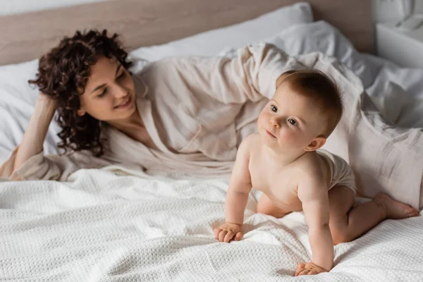 Mulher positiva com cabelo encaracolado sorrindo enquanto olha para a criança infantil rastejando na cama — Fotografia de Stock