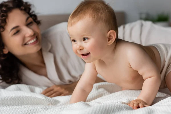 Mujer alegre con el pelo rizado sonriendo mientras que mira niño que se arrastra en la cama - foto de stock
