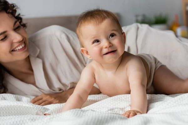 Joyful mother with curly hair smiling while looking at infant baby crawling on bed — Stock Photo