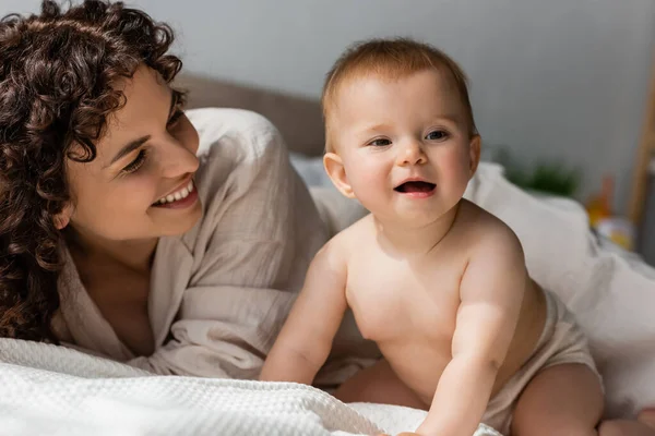 Madre con i capelli ricci sorridenti mentre guarda felice bambino infantile strisciare sul letto — Foto stock