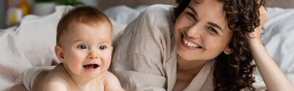 Happy woman with curly hair smiling while looking at camera near excited infant baby, banner — Stock Photo