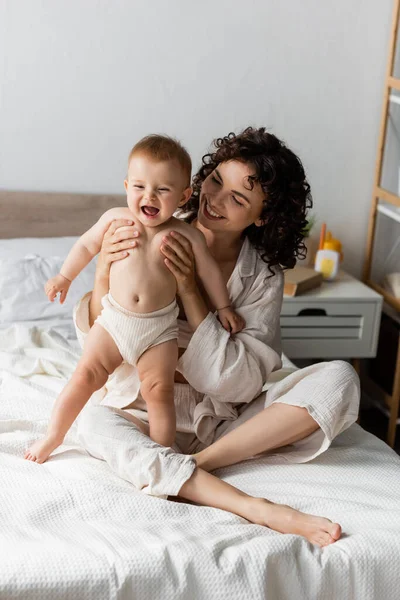 Mujer feliz con el pelo rizado sonriendo mientras sostiene en brazos bebé excitado - foto de stock
