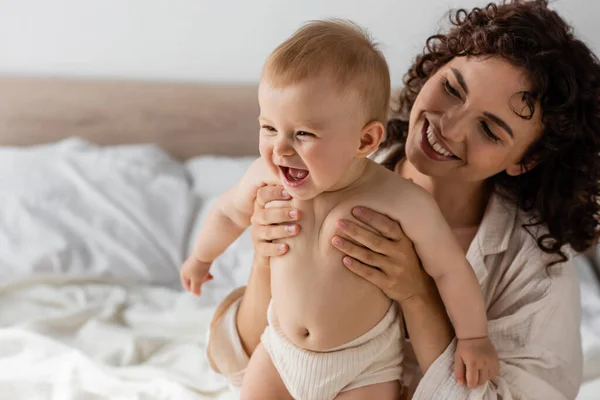 Cheerful woman with curly hair smiling while holding in arms excited infant baby — Stock Photo