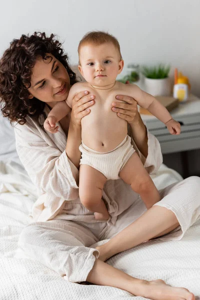 Pleased woman with curly hair smiling while holding in arms infant baby — Stock Photo
