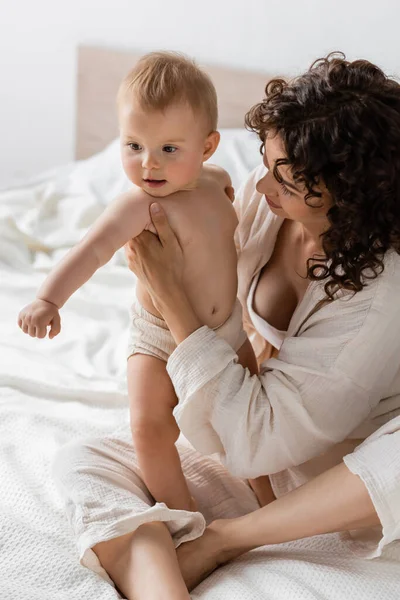 Mujer con el pelo rizado sentado en ropa de salón en la cama y sosteniendo en brazos hija bebé - foto de stock