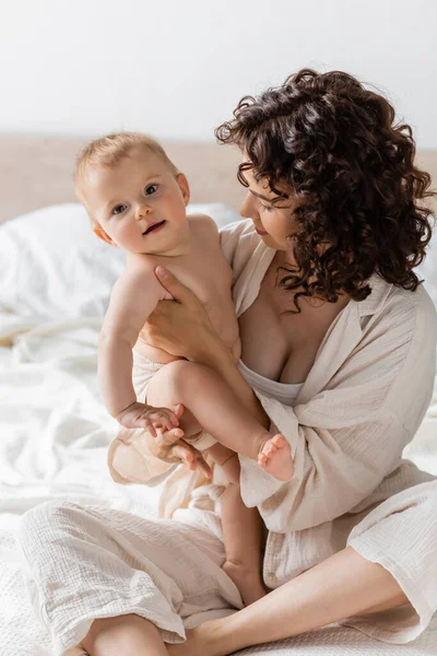 Mujer con el pelo rizado sentado en ropa de salón en la cama y sosteniendo en brazos feliz bebé hija - foto de stock