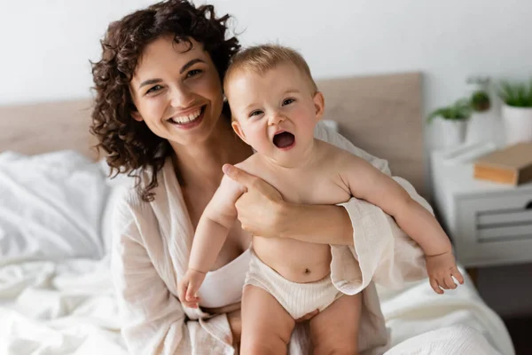 Mulher alegre em loungewear segurando nos braços bebê filha com boca aberta — Fotografia de Stock