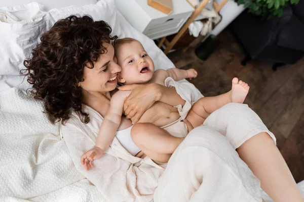 Ansicht von oben: freudige Frau in Loungewear, die ihre Tochter im Arm hält und auf dem Bett liegt — Stockfoto