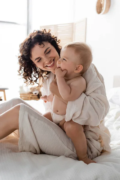 Fröhliche Frau in Loungewear, die ihre kleine Tochter im Arm hält und auf dem Bett sitzt — Stockfoto