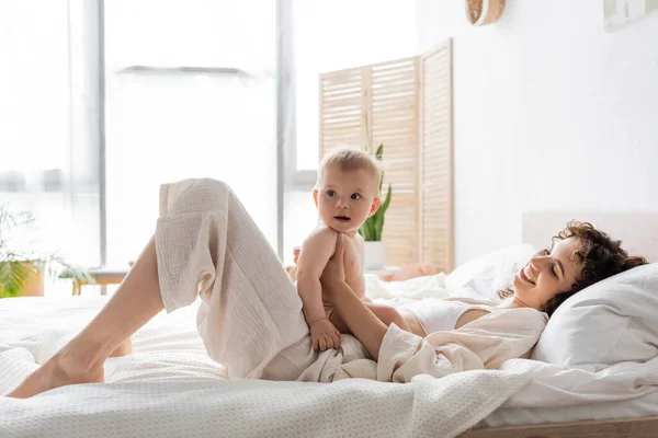 Mujer feliz en ropa de salón acostada en la cama y sosteniendo a la hija bebé - foto de stock