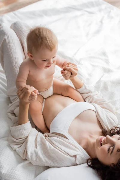 Blick von oben auf eine glückliche Frau in Loungewear, die auf dem Bett liegt und die Hände ihrer kleinen Tochter hält — Stockfoto