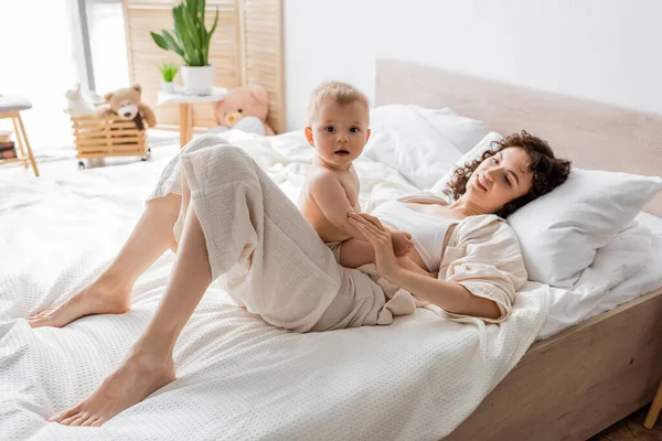 Mujer feliz en ropa de salón acostado en la cama y mirando curiosa hija lactante - foto de stock